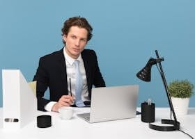 A focused businessman in a suit works at his tidy desk with a laptop, plant, and lamp.