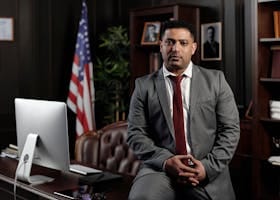 A professional businessman in a suit sitting in an office setting, with the American flag in the background.