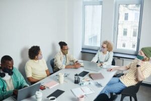 Office meeting with diverse colleagues wearing face masks, discussing projects in a modern workspace.