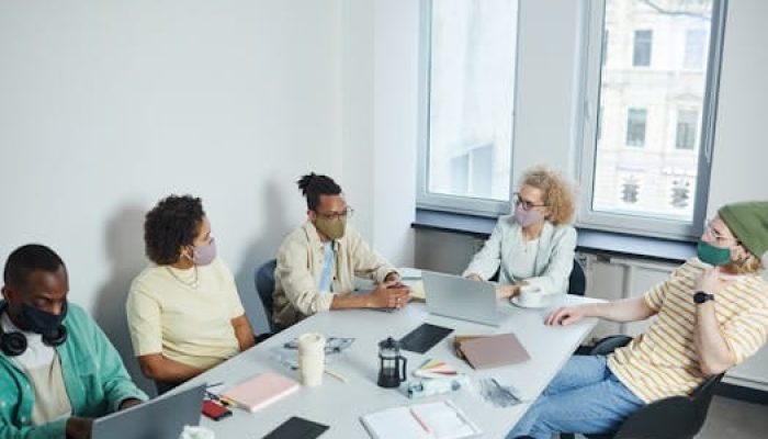 Office meeting with diverse colleagues wearing face masks, discussing projects in a modern workspace.